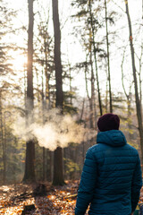 A man in a blue jacket stands in a serene forest surrounded by tall trees and soft sunlight shining...