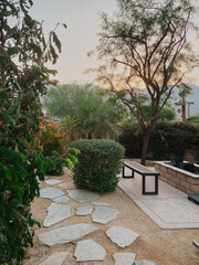 Sunrise desert backyard garden featuring an irregular flagstone path, modern wooden bench, built-in fire pit and lush desert plants with mountain backdrop in a tranquil outdoor living space