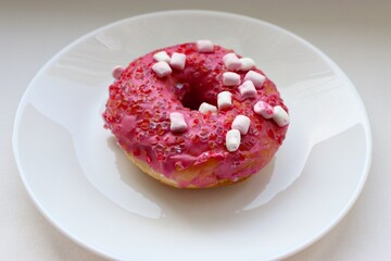 Glazed pink doughnut with marshmallows and colored sprinkles on a white plate, close-up of the dessert on a light background