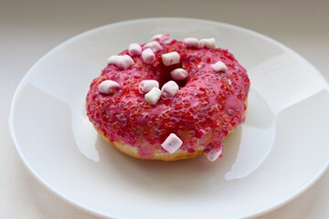 Glazed pink doughnut with marshmallows and colored sprinkles on a white plate, close-up of the dessert on a light background