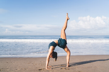 Sporty young woman performs graceful yoga pose with raised leg on sunny beach, demonstrating strength, balance, and digital focus through unplugged physical mindfulness.