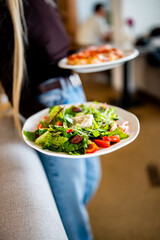 A person holds two plates—one with a fresh salad of greens, tomatoes, olives, and cheese, and another with pizza. Casual dining scene with a blurred background.