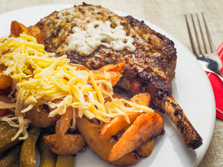 A plate of food with a premium pork chop in the bone, wedges potatoes, and tomatoes and cheddar cheese. The plate is set on a table with a fork and a knife. Fine tasty dinner with high quality meat.
