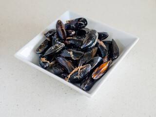 A white bowl filled with small black and orange mussels. The bowl is placed on a white countertop. Fine seafood product with delicate flavor and world wide production.