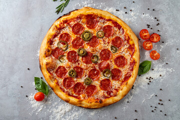 A delectable pepperoni pizza topped with spicy jalapenos sits center stage on a gray surface. Fresh basil leaves and vibrant cherry tomatoes enhance the presentation, inviting a slice