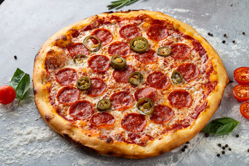 A perfectly baked pepperoni pizza topped with spicy jalapeños and fresh basil leaves rests on a textured gray surface. The vibrant colors create an inviting culinary experience