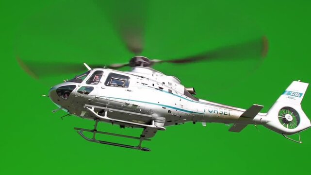 A white helicopter is filmed in flight against a solid green screen background.