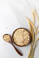 Top view of a wooden bowl with rolled oats and a wooden spoon, dried wheat stalks on a light marble background.