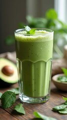 Green protein shake in clear blender cup on rustic table with spinach and avocado.
