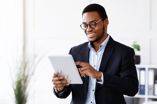 Fototapeta Modern technologies in business. Happy african american young businessman in suit using digital tablet in office, working online, reading marketing report, getting ready for presentation, copy space