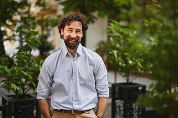 A man with curly hair and a beard stands in a vibrant outdoor area filled with plants, dressed in a light blue shirt. He looks relaxed and ready for business.