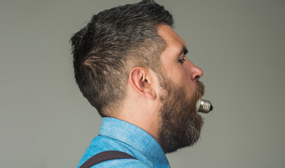 Obraz premium Bearded man with light bulb in mouth. Handsome man with stylish hairstyle in denim shirt eat light bulb lamp. Energy and electricity. Side view bearded man with electrical lightbulb in his mouth.