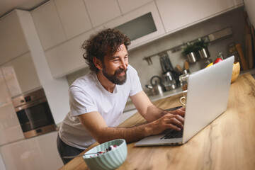 A man enjoys his morning by working on his laptop in a cozy kitchen, featuring a bowl of snacks and a cup of coffee on the table, emitting a productive and relaxed atmosphere.