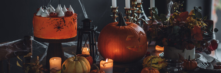 Homemade delicious chocolate cake with coloured orange cream and meringue ghosts, pumpkins for Halloween home party. Elegant beautiful table arrangement with candles, chandelier, Jack-o-Lantern banner