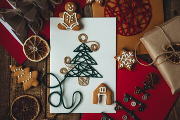 Flat lay. Preparation for a holiday season. Handmade children's greeting postcards with embroidered, origami fir tree, gingerbread cookies. Recyclable materials, eco friendly gift. Wooden background.