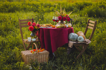 Beautiful decorated candlelight fall dinner, covered with a red tablecloth with flowers, pumpkins, candles, wine glasses. Outdoor in field near the forest in nature. Wedding banquet, sunset, evening