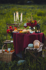 Beautiful decorated candlelight fall dinner, covered with a red tablecloth with flowers, pumpkins, candles, wine glasses. Outdoor in field near the forest in nature. Wedding banquet, sunset, evening