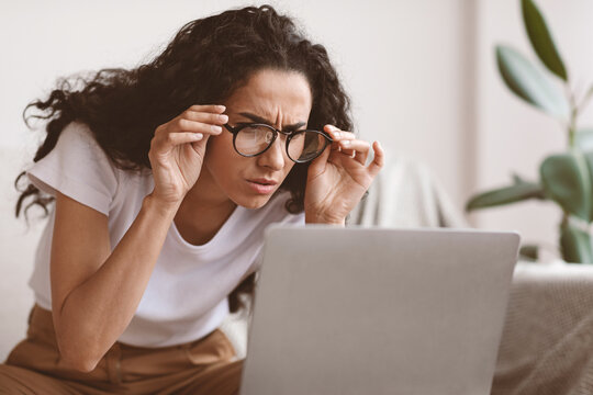 Young woman freelancer with bad eyesight using laptop, trying to work from home, copy space. Curly lady holding her glasses and squinting, looking at laptop screen, having vision troubles