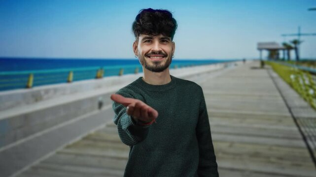 Young man extends hand invitingly on a seaside promenade with ocean background, showcasing his friendly demeanor and stylish appearance amidst a serene outdoor setting.