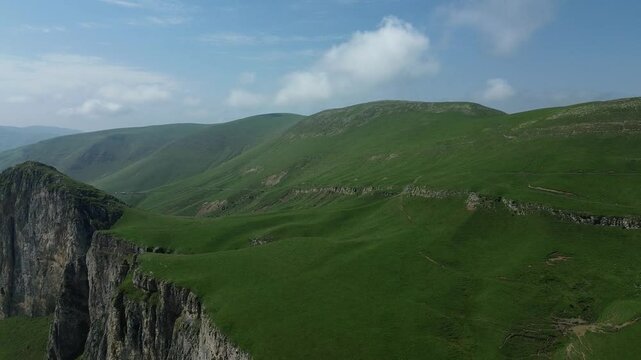 Beautiful panoramic view of green mountains with rocky formations, covered in lush grass, captured by drone flying backward against a clear blue sky.
