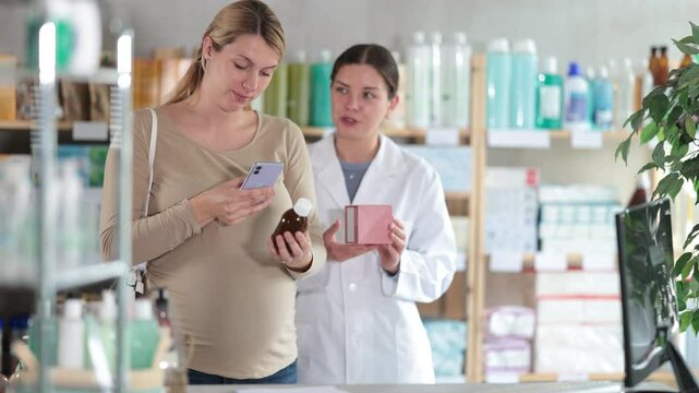 Modern pregnant woman using phone to scan QR code on bottle of syrup and check ingredients or reviews online, while polite pharmacist preparing to offer professional advice or alternative in pharmacy
