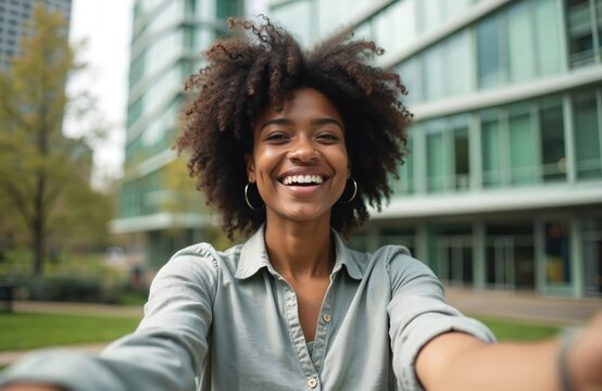 Happy black woman takes selfie photo outdoors. Smiling girl with afro hairstyle and toothy smile in city park. She remembers moments, takes profile pic for social media.