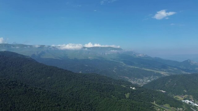 Drone flight over mountains covered with dense forests, steep cliffs visible on the horizon, blue sky with clouds resting on the mountains.

