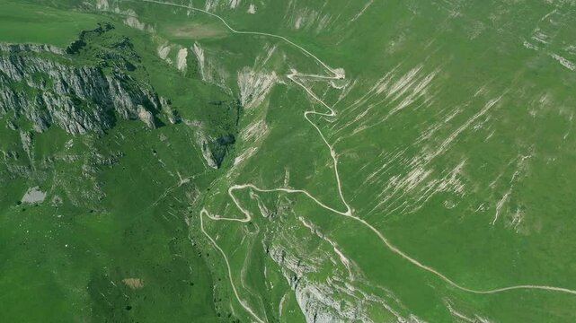 Drone footage of mountainous terrain covered with lush grass and forests, featuring clear mountain roads and steep rocky cliffs. Clouds rest on the mountains with a blue sky on the horizon.
