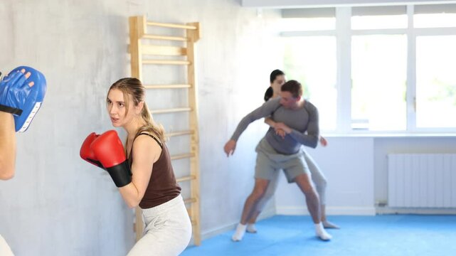 Sportive young female practitioner of boxing courses applying kicks on hitting mitts during workout session