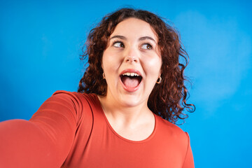 Curvy model with curly hair taking a selfie with a surprised expression against a vibrant blue background