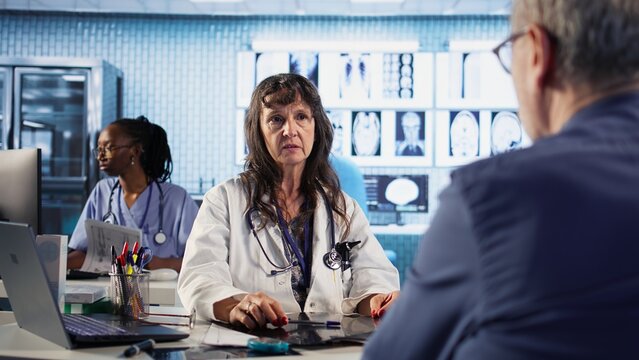 Female general practitioner giving bad worrying news to elderly man, reading the x ray results and finding a dangerous diagnosis. Scan interpretation for a concerned senior patient. Camera A. - Powered by Adobe