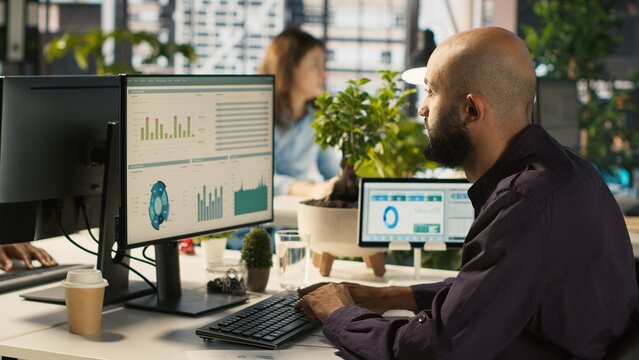 Employee editing documents on desktop computer at office desk, updating reports and files. Worker analyzing data charts on computer screen at desk, interpreting statistical information, camera A