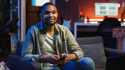 Smiling young adult chatting with friends over texts on mobile phone, sitting on the couch and...