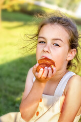 Little curly girl in a light summer sundress eating a croissant. Summer day in a park or garden. Close-up, looking ahead. Blurred background.