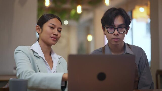 Young professional woman mentoring a barista in front of a laptop at a coffee shop. Female business coach or consultant teaching small business operations, technology usage.