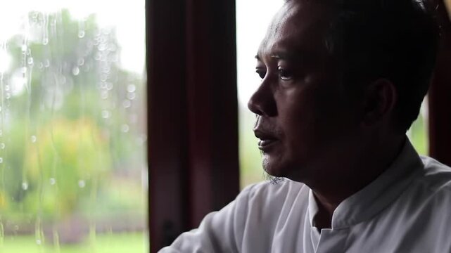  An elderly man sits by a window, gazing outside thoughtfully. Natural light highlights his silhouette, creating a reflective and calm mood.