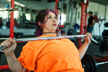 Beautiful, overweight Latina woman concentrating on lifting weights on an exercise machine at a gym. Wearing colorful clothes, hair, and retro makeup, she's positive. She's pushing herself.
