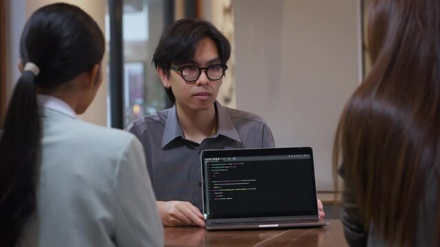 Young Asian software developer showing source code on a laptop screen to colleagues during a team discussion or project meeting in a modern workspace. Scene portrays teamwork in tech, software develop