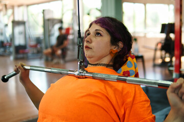 Beautiful, overweight Latina woman concentrating on lifting weights on an exercise machine at a gym. Wearing colorful clothes, hair, and retro makeup, she's positive. She's pushing herself.
