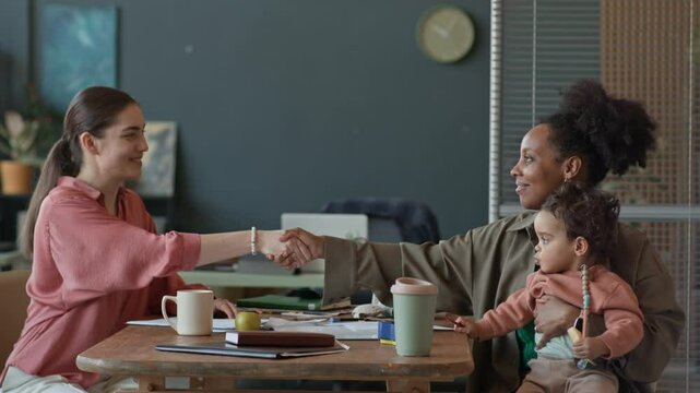 Side view of young African American mother with baby shaking hand of female social worker at table in office