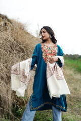 A female Kashmiri model wearing a traditional pheran and shawl, posing for the artisanal brand Kaar Kash, with a richly embroidered curtain as a backdrop in a serene outdoor setting.

