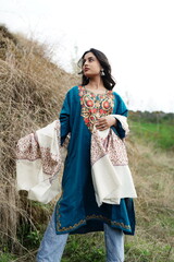 A female Kashmiri model wearing a traditional pheran and shawl, posing for the artisanal brand Kaar Kash, with a richly embroidered curtain as a backdrop in a serene outdoor setting.

