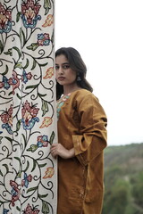 A female Kashmiri model wearing a traditional pheran and shawl, posing for the artisanal brand Kaar Kash, with a richly embroidered curtain as a backdrop in a serene outdoor setting.

