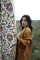 A female Kashmiri model wearing a traditional pheran and shawl, posing for the artisanal brand Kaar Kash, with a richly embroidered curtain as a backdrop in a serene outdoor setting.

