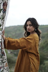 A female Kashmiri model wearing a traditional pheran and shawl, posing for the artisanal brand Kaar Kash, with a richly embroidered curtain as a backdrop in a serene outdoor setting.

