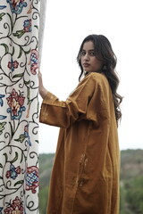 A female Kashmiri model wearing a traditional pheran and shawl, posing for the artisanal brand Kaar Kash, with a richly embroidered curtain as a backdrop in a serene outdoor setting.

