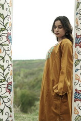 A female Kashmiri model wearing a traditional pheran and shawl, posing for the artisanal brand Kaar Kash, with a richly embroidered curtain as a backdrop in a serene outdoor setting.

