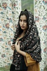 A female Kashmiri model wearing a traditional pheran and shawl, posing for the artisanal brand Kaar Kash, with a richly embroidered curtain as a backdrop in a serene outdoor setting.

