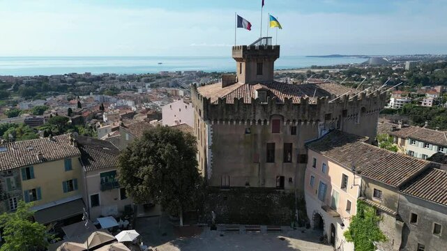 A historic stone castle with flags on top, surrounded by trees and buildings. Clear blue sky, medieval architecture, background visuals for vlogs or social media posts about France, travel and tourism