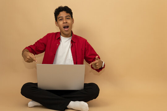 Young brown skinned Latino man sitting with a shocked expression pointing at his laptop with his hands on a light brown background with advertising space